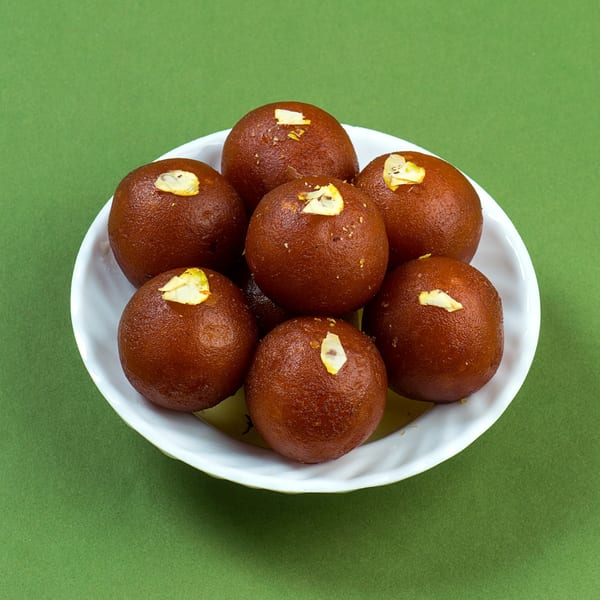 A white bowl filled with several brown gulab jamun, garnished with slivered nuts, on a green background.