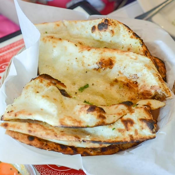 Basket of golden-brown Indian naan bread, some pieces folded, with visible charring and herbs.